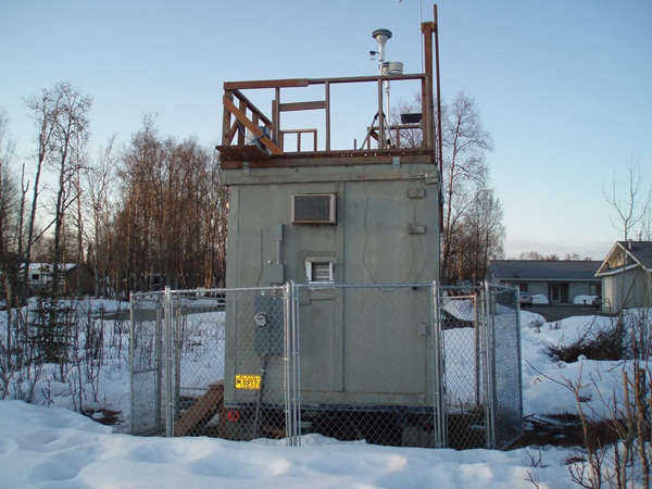 A wooden trailer surrounded by chain link fence, atop the trailer is a deck railing and on the deck there are two inlets for sampling air quality. Snow covers the ground around the trailer.